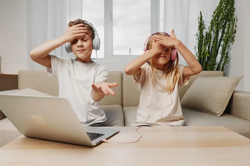 Two children with headphones sit on a sofa using a laptop, covering their eyes with hands in a bright living room. Concept of digital interaction and playful technology use.