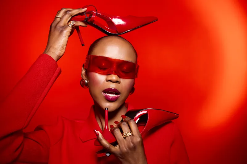 Stylish black woman with dark skin holds red high heels and matching lipstick wearing red sunglasses and coat. Bold fashion concept with vibrant red background and confident expression.