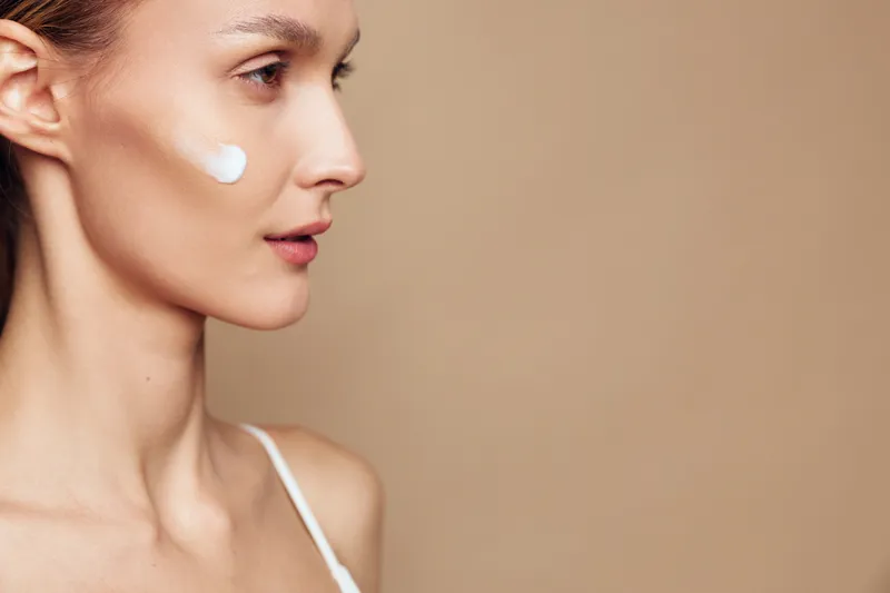 A close side profile of a young woman with a dollop of facial cream on her cheek, set against a warm beige background, illustrating skincare routine and calm beauty.