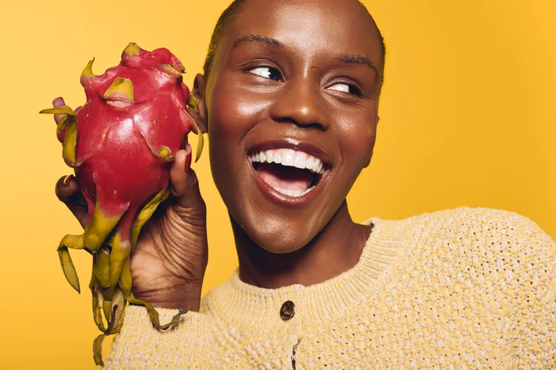 Smiling African woman with dark skin holds dragon fruit near her face against yellow background. Concept of healthy lifestyle, exotic fruit, nutrition, and joyful mood in studio.