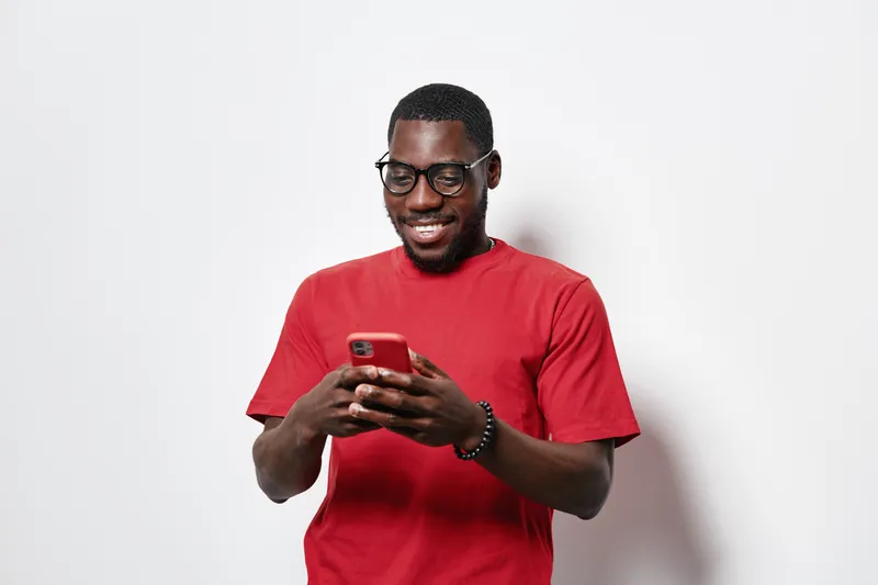 African American man with dark skin wearing glasses and red t shirt smiling while using smartphone for texting or social media on white background.