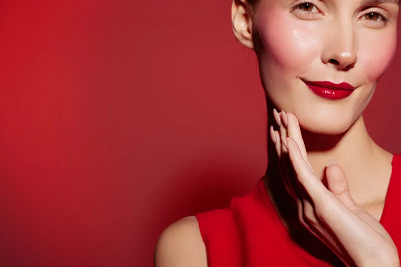 Close-up of a smiling woman with red lipstick and smooth skin touching her face against a red background. Concept of beauty, skincare, makeup, and confidence.