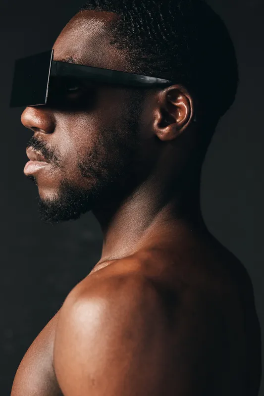 African man with dark skin wearing black sunglasses in creative conceptual visual style. Closeup portrait with bare shoulders and textured skin on dark background for artistic expression.