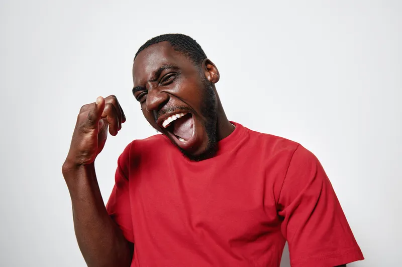 African American man with dark skin wearing red t shirt expressing excitement and joy with clenched fist and open mouth on white background. Emotion, celebration and happiness.