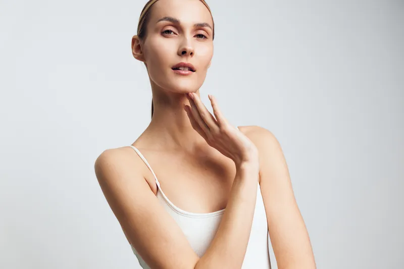 A young woman with clear skin poses with a gentle touch on her neck, wearing a white tank top against a plain light background. Concept for skincare, beauty, and natural wellness.