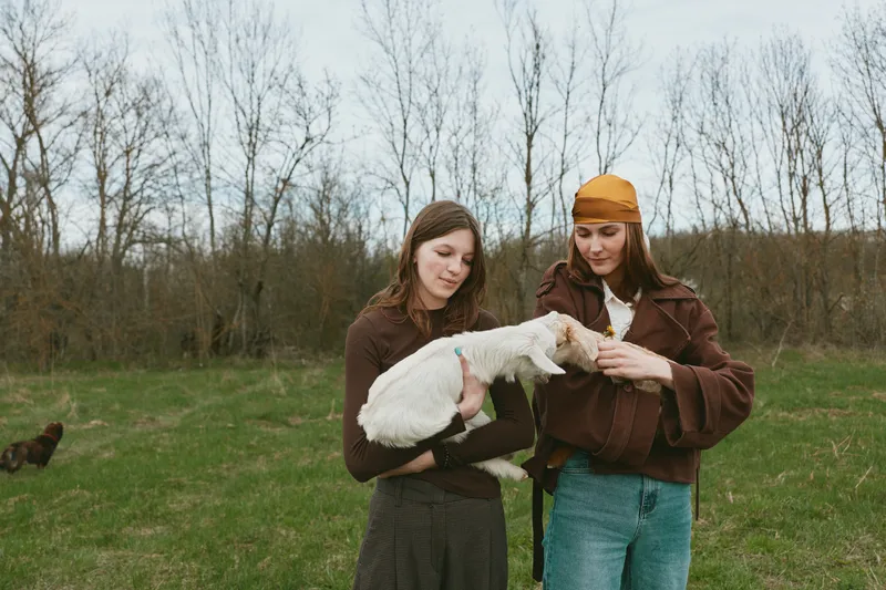 Cheerful woman outdoors holding a white goat kid in a grassy field, a calm moment highlighting a simple lifestyle among animals in a countryside setting