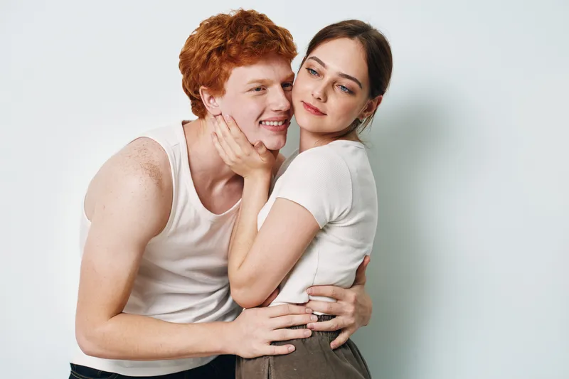 Young couple embracing with woman holding mans face tenderly and man smiling for love, affection, relationship and connection on light background.