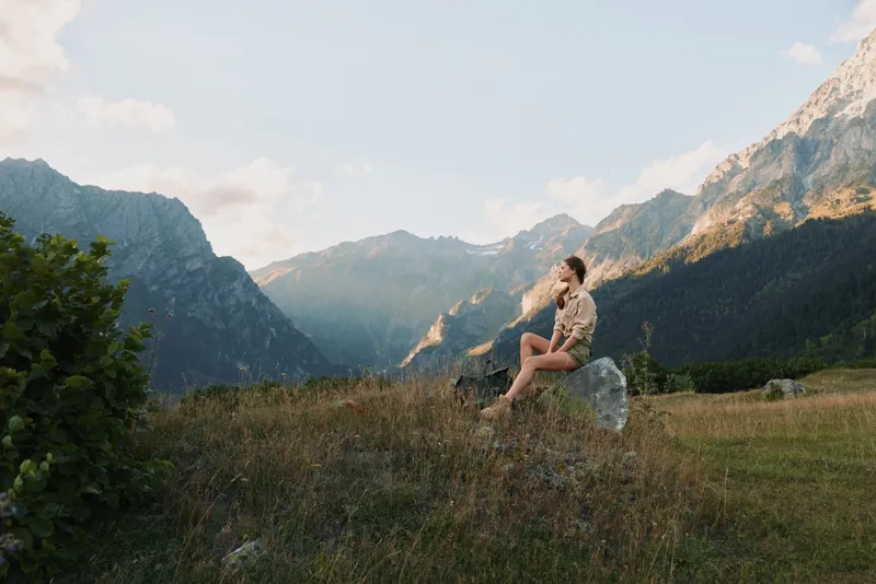 Young woman sits on a rock in a meadow with distant mountains, enjoying a peaceful sunny moment.