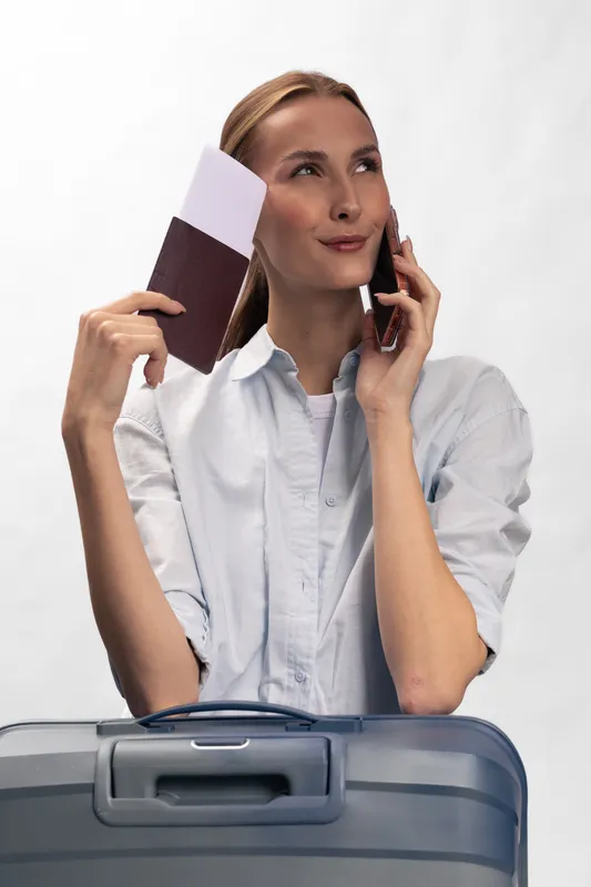Young woman in casual shirt holds passport and plane ticket while talking on phone, standing behind suitcase. Concept of travel planning, vacation, and trip preparation.
