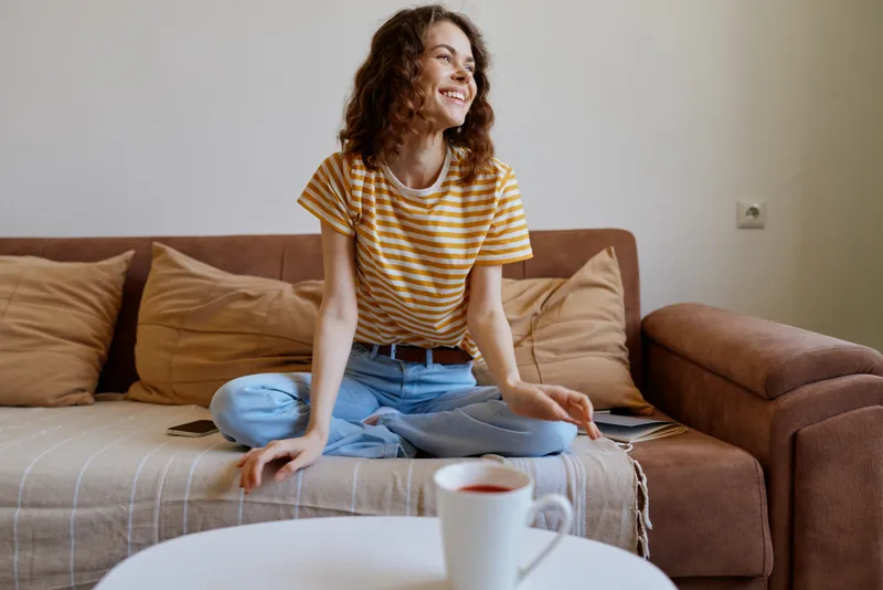 A smiling young woman with curly hair sits cross-legged on a sofa in a bright living room, wearing a striped tee and jeans, coffee mug on the table nearby.