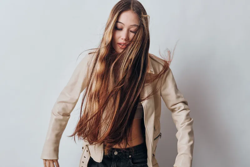 Woman with long brown hair wearing beige jacket and black pants posing against plain light background in studio, casual style and natural expression indoors.
