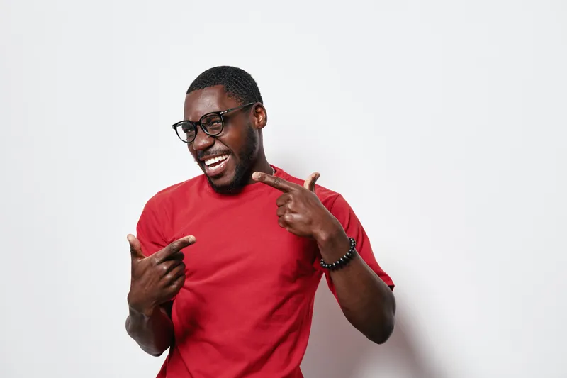 African American man with dark skin wearing glasses and red t shirt smiling and pointing fingers at himself on white background. Happy confident black male portrait with casual style.