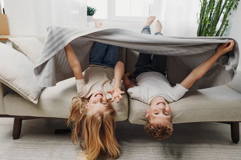 Two happy children lie upside down on a sofa under a gray blanket in a bright living room. Playful mood, family bonding, cozy home atmosphere, and childhood joy captured in natural light.
