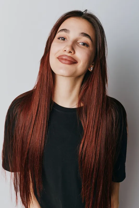 Young woman with long red hair and natural makeup smiling confidently in black t shirt against plain white background. Portrait of happy female with casual style and earrings.
