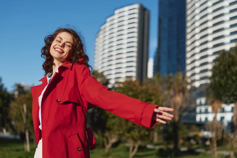 Happy woman outdoors smiling with eyes closed wearing red coat urban park sunny day modern buildings background positive mood