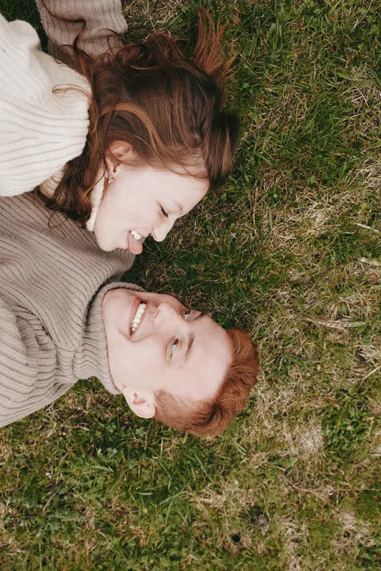 Young couple lies on green grass smiling and looking at each other enjoying a relaxed and joyful moment in casual sweaters outdoors in natural light