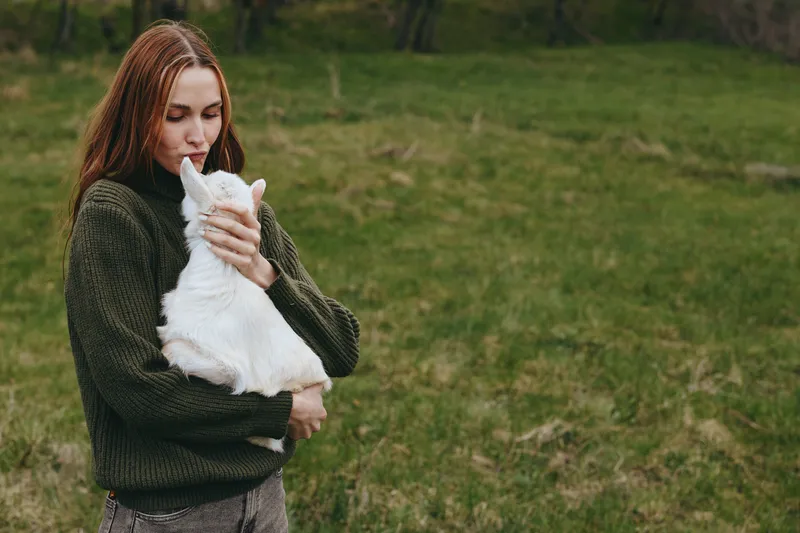 A woman in a dark sweater cradles a white goat kid in an open field, sharing a tender outdoor moment that highlights rural life, companionship, and gentle bonding.