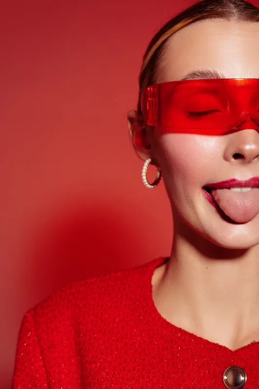 Young woman in red sweater and futuristic red glasses sticks out tongue with eyes closed. Studio portrait on red background. Concept of playful attitude, fashion, and modern style.