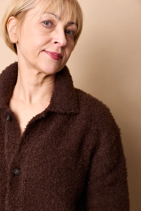 Elderly woman portrait, short blond hair, warm smile, closeup face, dark sweater, calm expression, indoor soft light, beige background