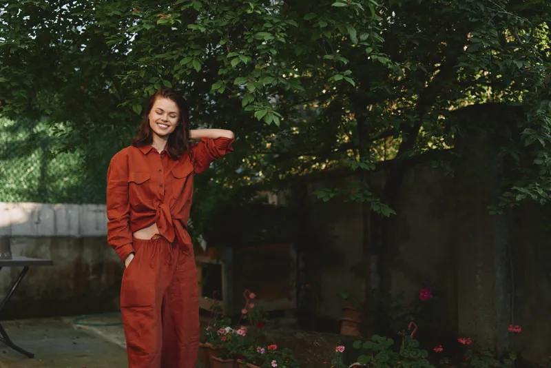Cheerful woman outdoors in orange outfit, candid pose, sunlit moment, relaxed posture, lush garden background, natural colors, breeze