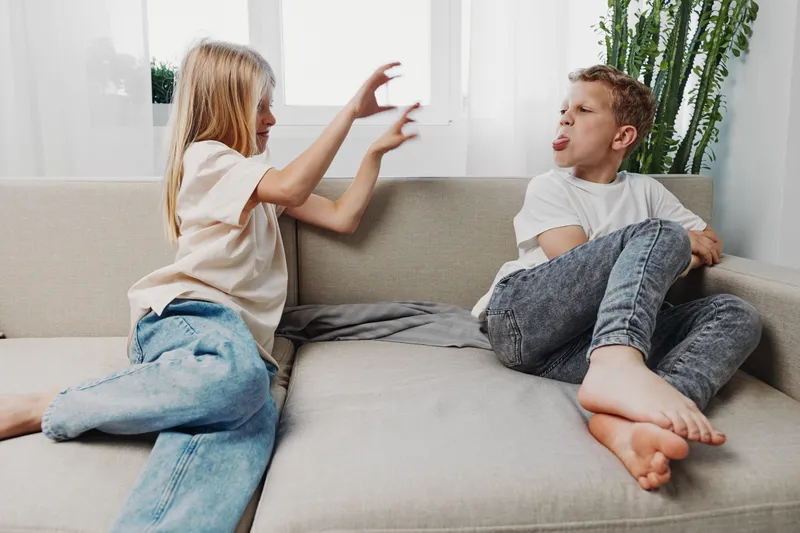 Two children sit on a couch in a bright room. The girl playfully reaches toward the boy who reacts with a funny face. Concept of sibling interaction, childhood fun, and family moments.
