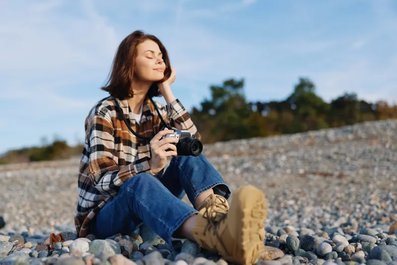 Woman photographer seated on a pebbled shore, holding a camera, wearing a plaid shirt and rugged boots, enjoying sunny weather and a peaceful outdoor moment in nature.