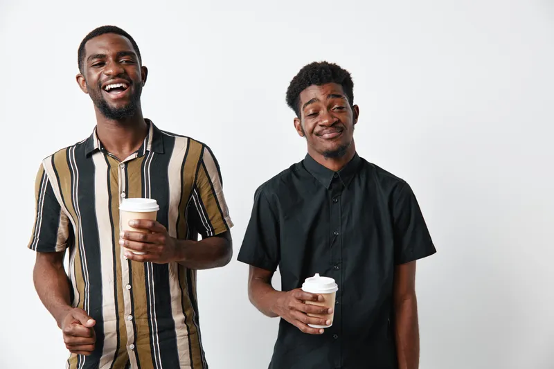 Two young African American men with dark skin holding coffee cups smiling and standing against white background in casual striped and black shirts indoors.