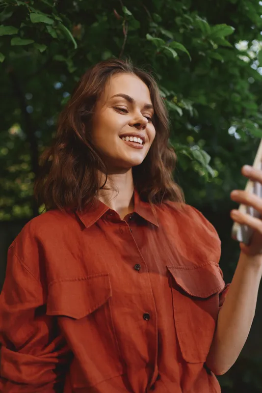 Young woman with shoulder length brown hair smiles while looking at her phone outdoors, surrounded by lush green foliage, wearing an orange casual shirt and enjoying a relaxed moment