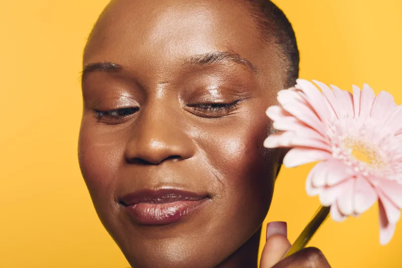 Close-up of a happy African woman with dark skin holding a pink flower near her face on a yellow background. Concept of natural beauty, skincare, wellness, and positive mood.