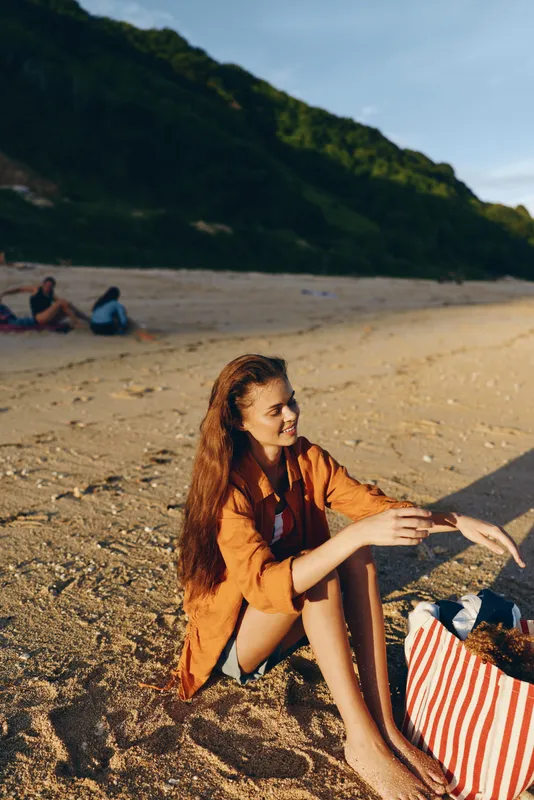 Young woman with long red hair sits on sandy beach wearing orange shirt and denim shorts while reaching into striped bag under warm sunlight with hills in background.