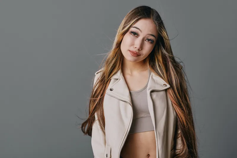 Young woman with long hair wearing beige jacket and crop top posing on gray background. Fashion model with casual style and makeup looking at camera.