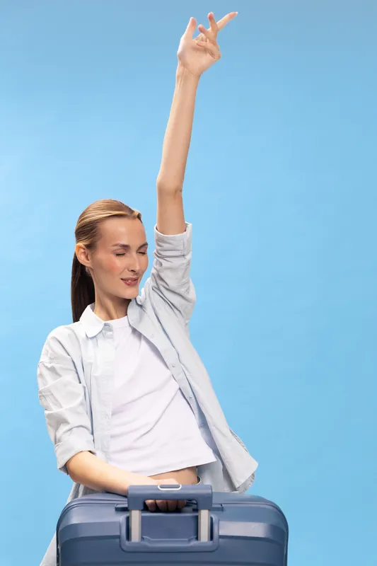Happy woman with suitcase raising arm and smiling with closed eyes on blue background. Travel concept, vacation mood, casual outfit, freedom and holiday spirit captured in studio.