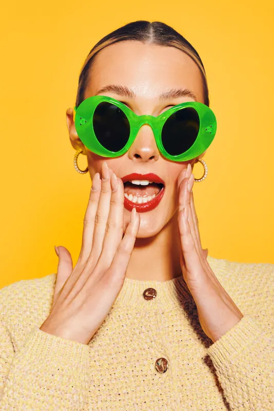 Young woman with bright green sunglasses and red lipstick expresses surprise with hands near face. Yellow background and beige sweater create vibrant, playful fashion and beauty concept.