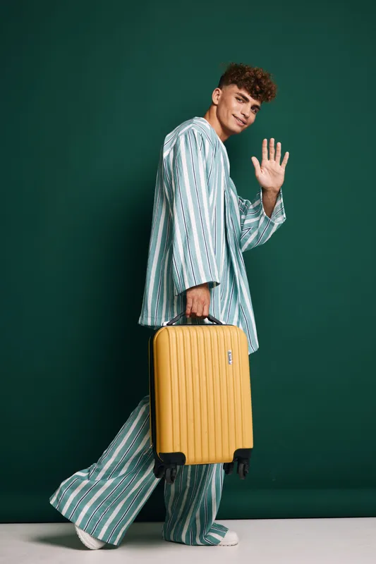 Young man in striped pajamas walking with yellow suitcase waving hand against green background for travel, vacation and casual style with luggage and smile.