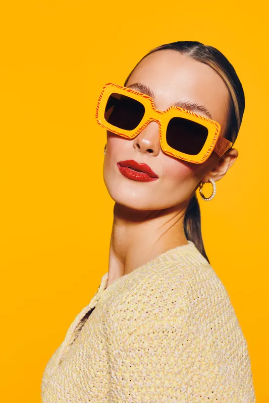 Stylish woman with slicked back hair wearing oversized orange sunglasses and red lipstick poses against bright yellow background. Fashion concept with bold colors and confident attitude.