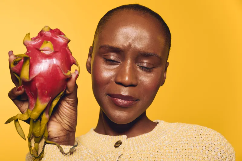 Closeup of dark skinned African woman holding exotic dragon fruit near face with closed eyes. Yellow background and beige sweater create warm and natural concept of healthy lifestyle and fruit.