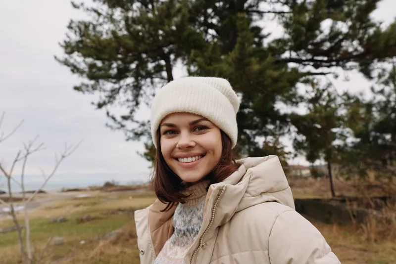 Woman in a cozy beige jacket and knit beanie outdoors, smiling with a scenic field and distant water behind her, amid trees and crisp air.