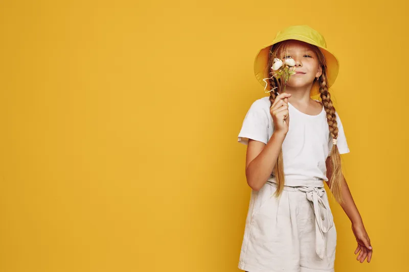 Smiling girl with two braids wearing a yellow hat holds white flowers on a yellow background. Studio portrait of a happy child in casual clothes.
