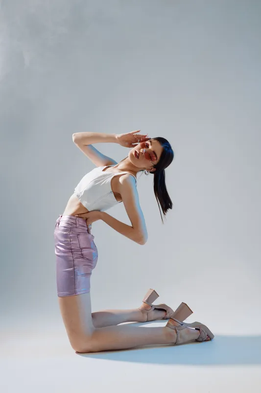 Young woman in a white top and shiny purple skirt poses kneeling with hand on forehead. Studio shot with soft lighting. Concept of woman beauty fashion and elegance.