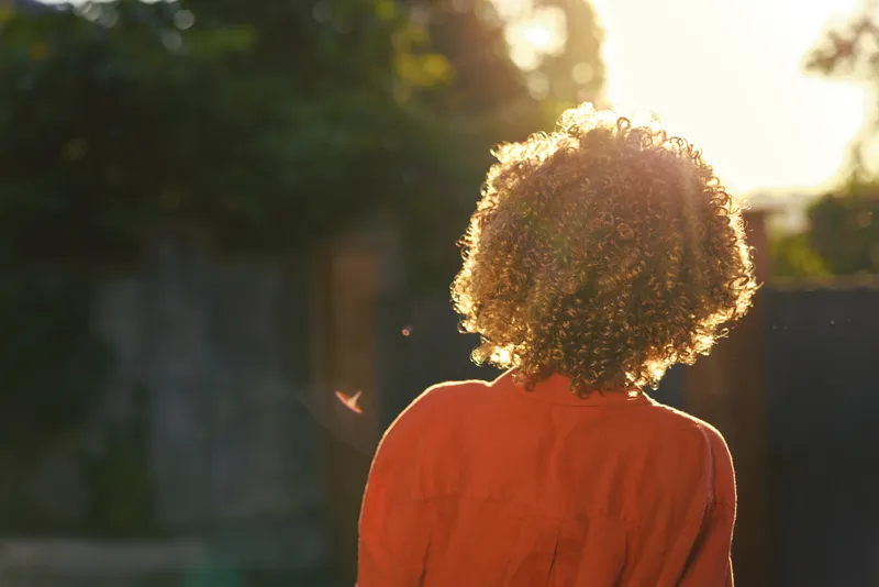A woman seen from behind in an orange top stands outdoors at sunset, presenting a calm lifestyle moment. Warm light rims curly hair, conveying serenity and simple, reflective everyday life