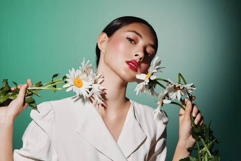 Fashionable woman with glowing skin and red lips holds blooming daisies near her face on green background. Concept of beauty, creative fashion, and natural elegance.