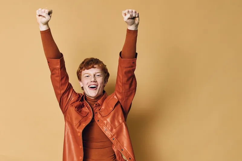 Young man with red hair in orange jacket raising hands with excitement and smile on beige background for happiness and celebration.