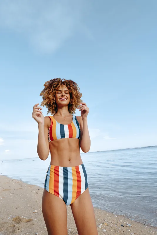 Woman with curly hair wearing striped swimsuit smiling on a beach by the ocean, summer vacation mood, soft wind, bright natural light, relaxed happy pose, blue sky background