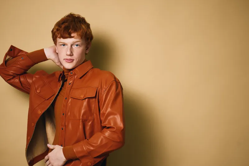 Young man with red hair wearing orange leather jacket standing against beige background posing with hand behind head looking at camera with neutral expression and casual style.
