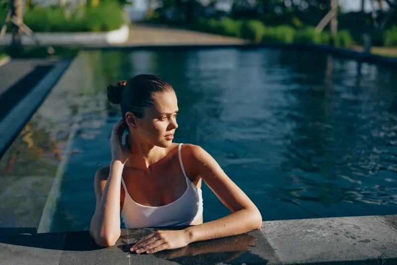 Young woman in a white tank top leans on the pool edge, sunlight on water creates a calm inviting mood. A lifestyle moment focused on relaxation, wellness, and leisure.
