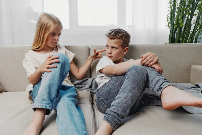 Two children sitting on sofa indoors, a girl teasing a boy with playful gesture while both wear casual clothes in bright living room with natural light and plants in background.