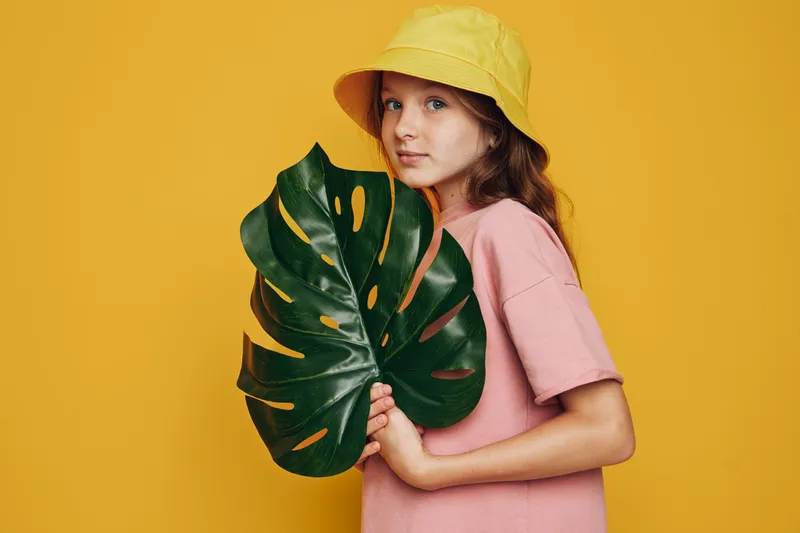 Young girl in a yellow hat and pink shirt holds a large green monstera leaf on a solid yellow background. Studio portrait with natural lighting.