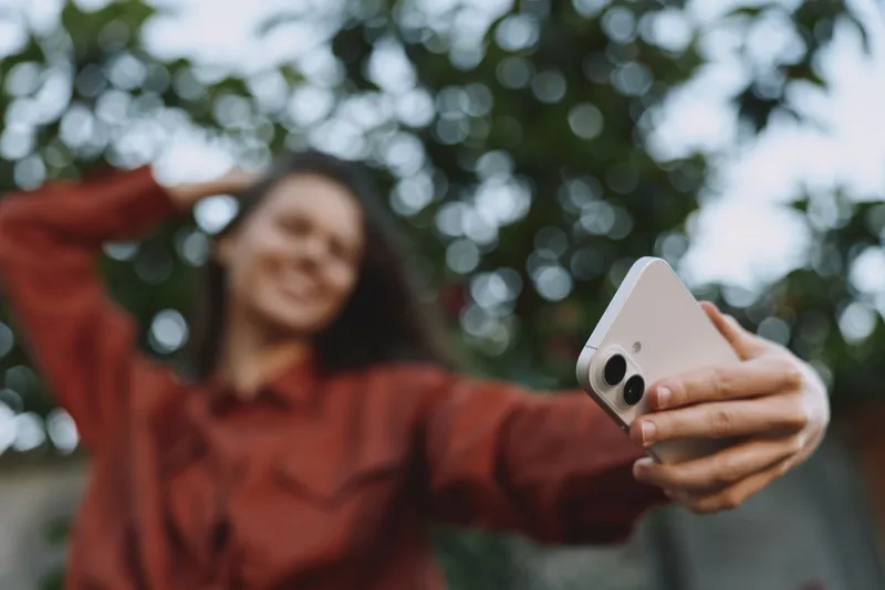 A smiling woman outdoors takes a selfie with a smartphone, embracing a casual lifestyle. Natural light frames green trees in the background, highlighting everyday joy and self expression