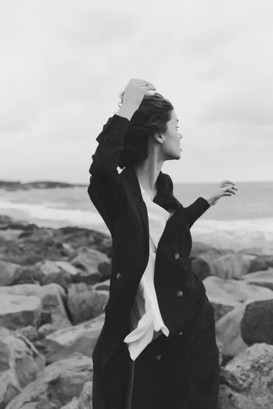 Black and white seascape with a windblown woman in a long coat standing on a rocky shore, capturing motion, mood and ocean breeze against the dramatic horizon, timeless coastal atmosphere and breeze