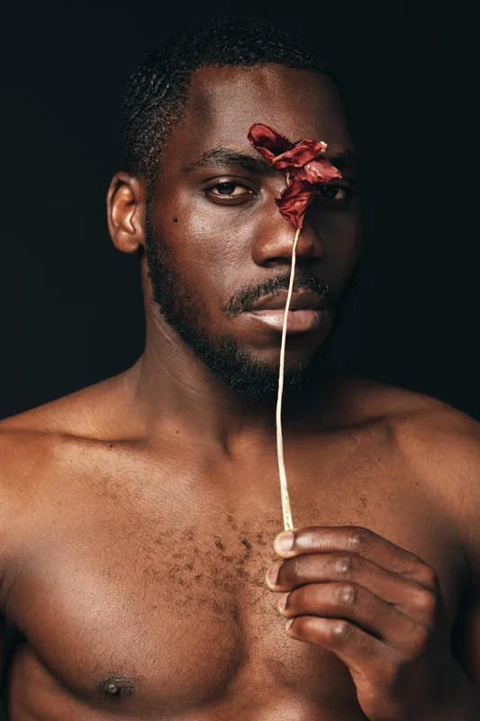 Creative conceptual portrait of African man holding dried flower in front of face with serious expression, bare chest and dark background for artistic visual style and emotion.
